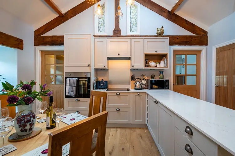 A bright, modern kitchen in Silverdale with white cupboards, a large island, wooden beams, and vaulted ceiling—perfect for Holiday Accommodation. Dining table with flowers and stylish place settings in the foreground enhances the cottage charm.