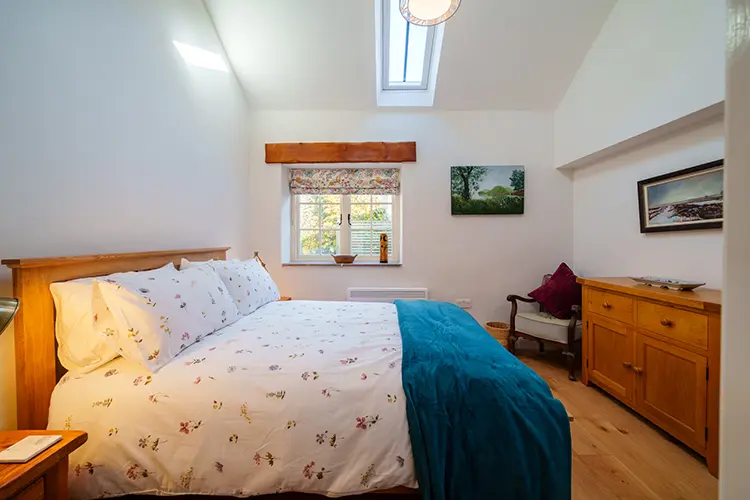 A cosy bedroom in Lancashire with a large bed, white floral bedding, and a teal blanket. This charming holiday accommodation features wooden furniture, artwork on the walls, natural light from a window, and a skylight above.