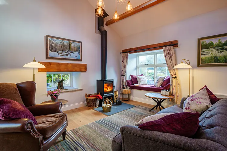 Cosy living room with a wood-burning stove, armchair, sofa with purple cushions, window seat, coffee table, and warm lighting. Large windows and wall art add charm—perfect for relaxing holiday cottages in Lancashire near scenic Morecambe Bay.