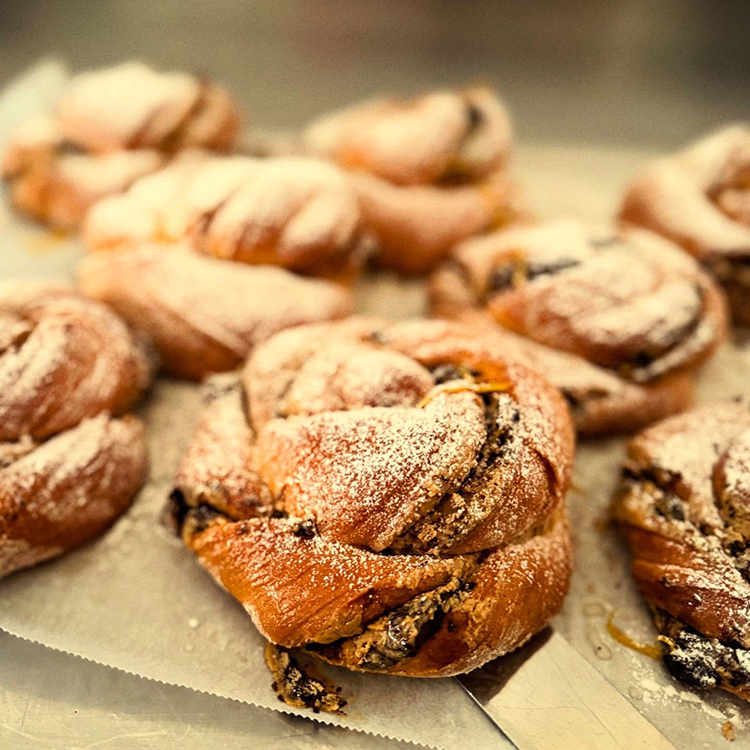 A close-up of several twisted pastries dusted with icing sugar, arranged on a baking tray. The pastries have a golden-brown crust and visible swirls of dark filling.