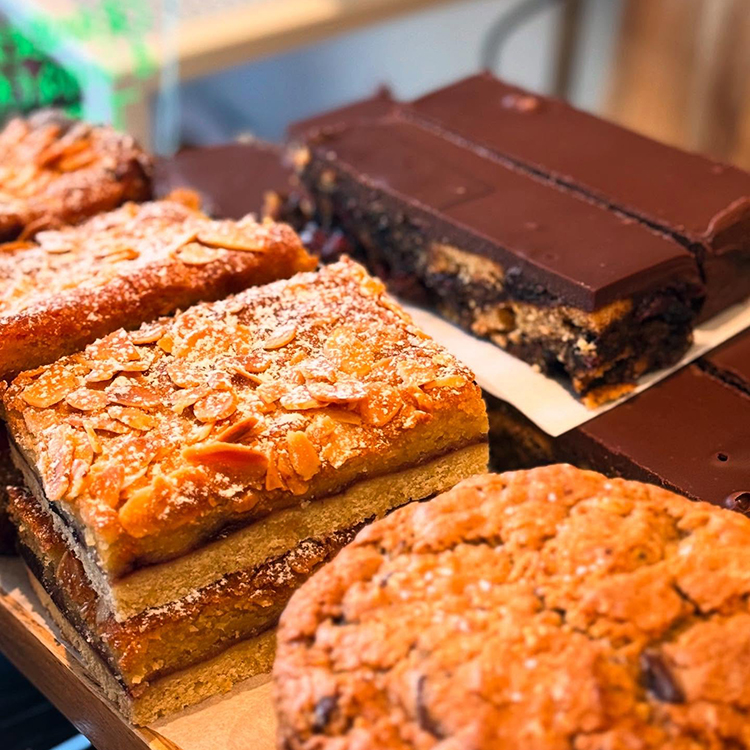 Close-up of assorted baked goods, including almond-topped bars, thick chocolate brownies with a layered filling, and a large, round oat biscuit on a wooden surface.