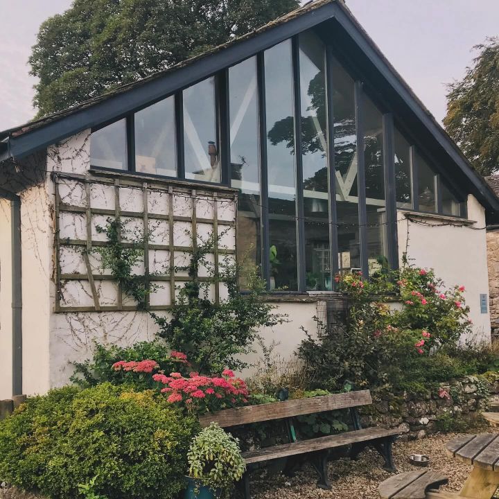 A building with large glass windows and a slanted roof is surrounded by lush greenery, flowering plants, and bushes. Wooden benches are in front, and trees are visible in the background.